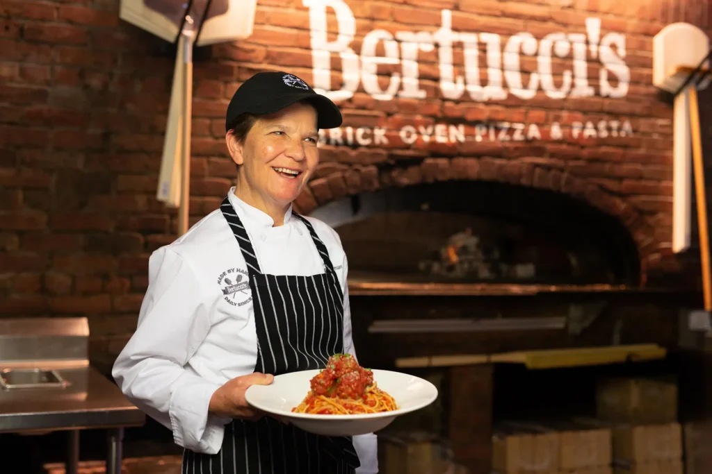 Bertucci’s chef smiling while holding a plate of spaghetti and meatballs in front of a brick oven at a Bertucci’s restaurant.