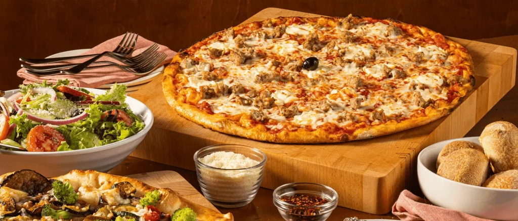 A spread of Bertucci's food with a sporkie pizza, roasted vegetable pizza, insalata salad, and rolls with crushed red pepper and parmesan cheese