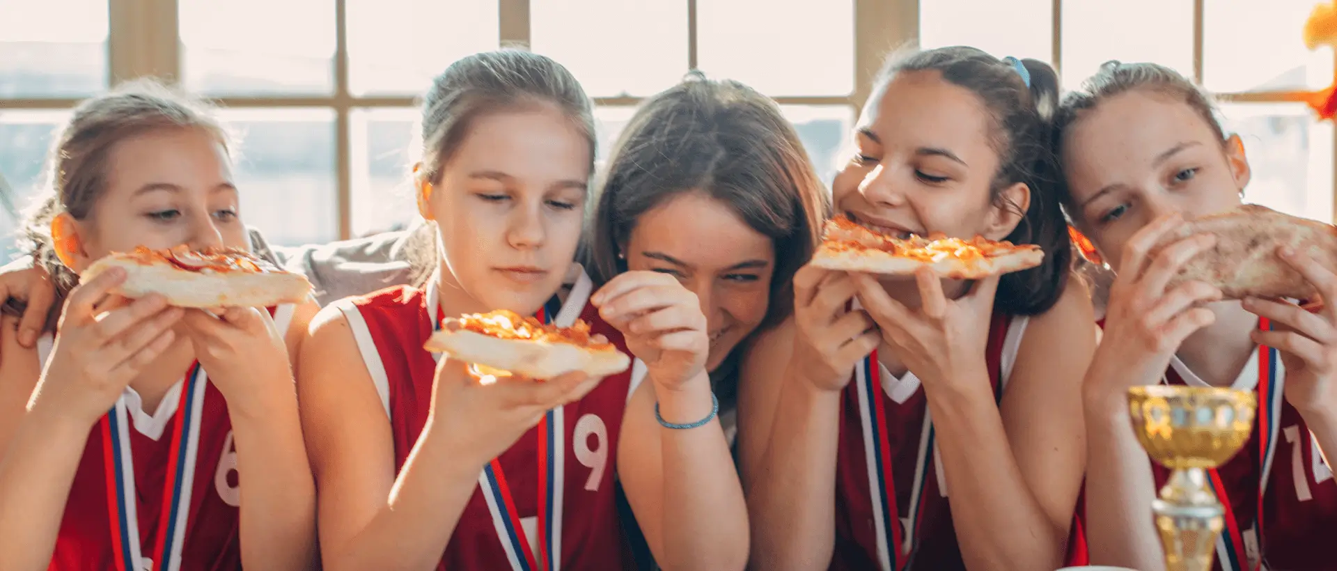 A group of kids in sports uniforms eating pizza while wearing medals